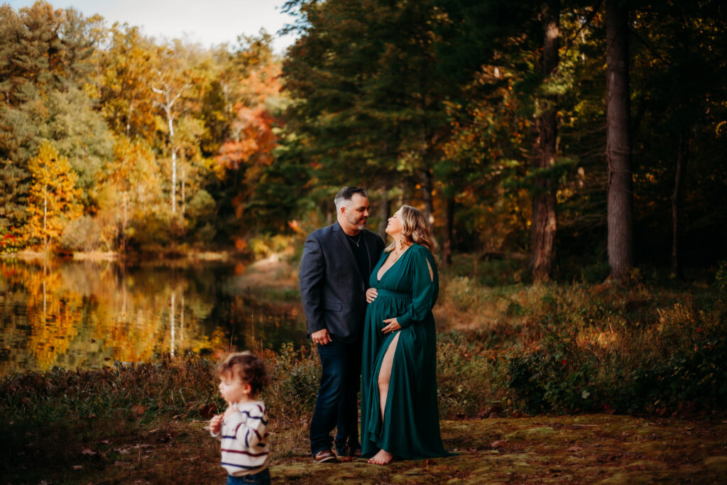 husband and wife next to each other in lush fall forest while little boy runs around 